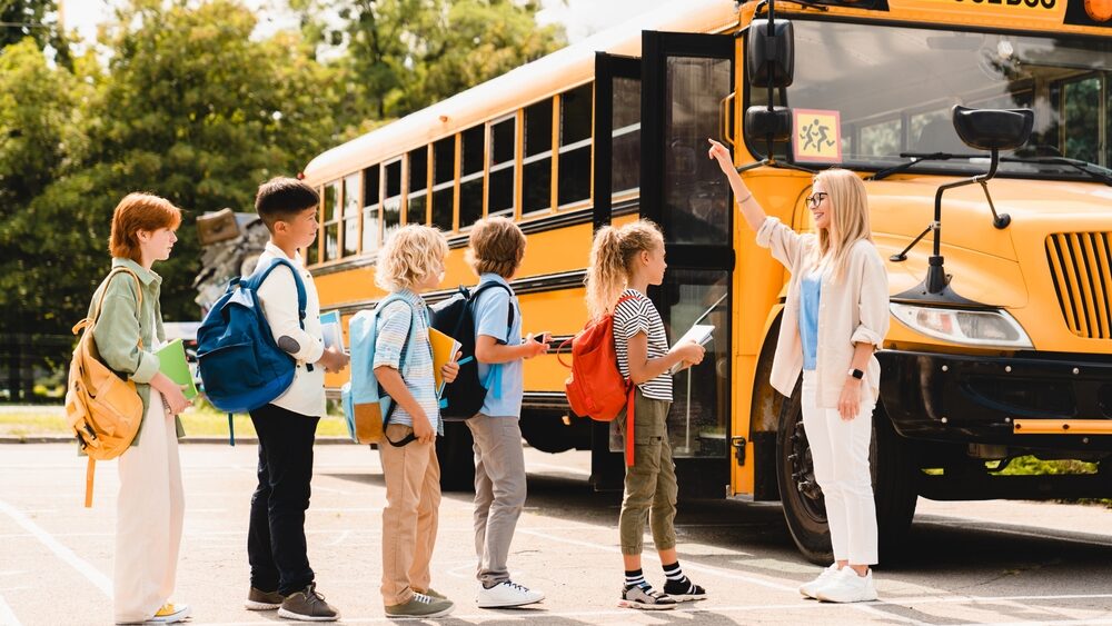 Young teacher counting group of kids pupils schoolchildren before boarding school bus before lessons. Welcome back to school after summer holidays! New educational year semester.